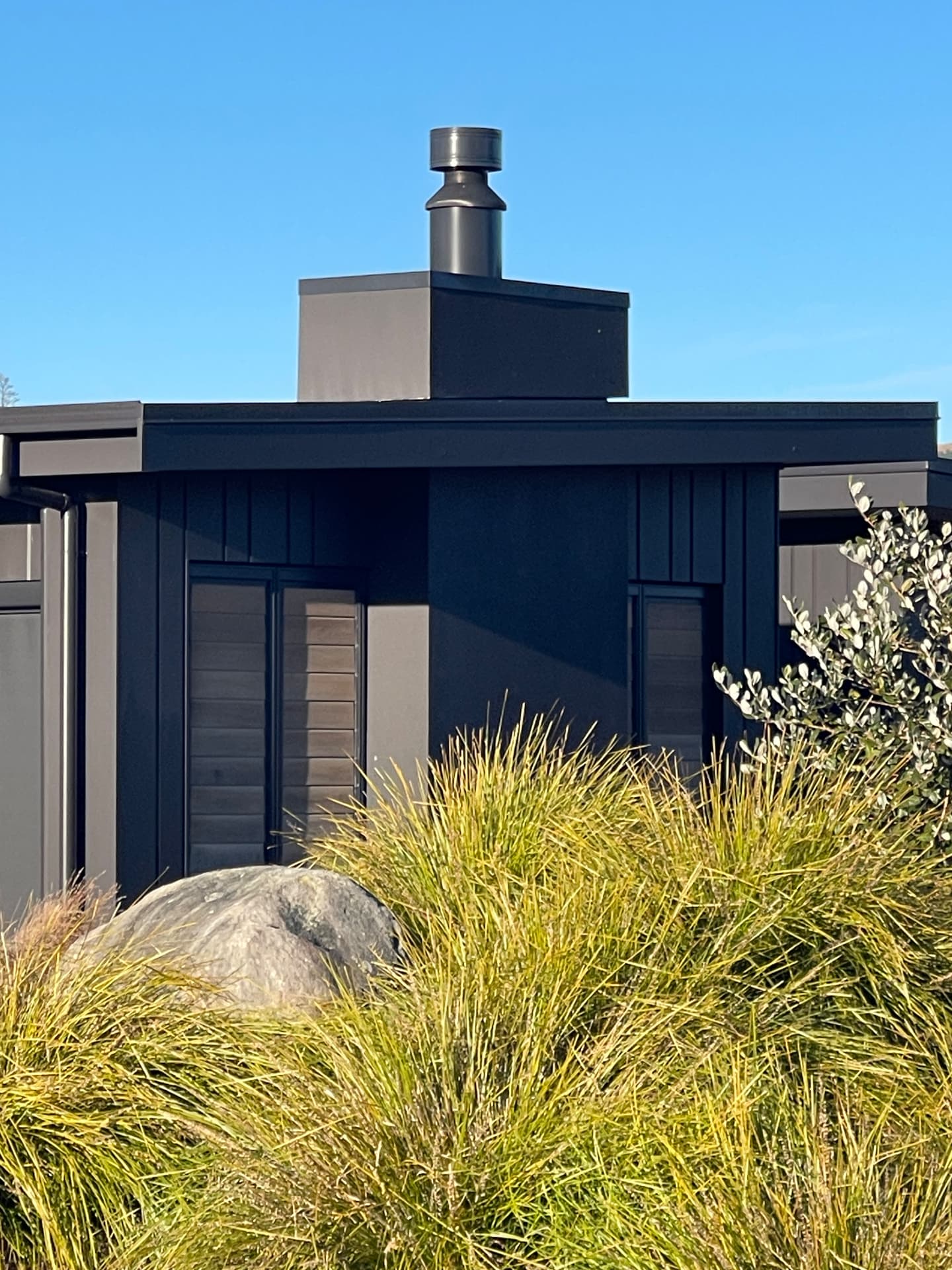 Black board-and-batten house with a squared chimney above golden tussock and a boulder